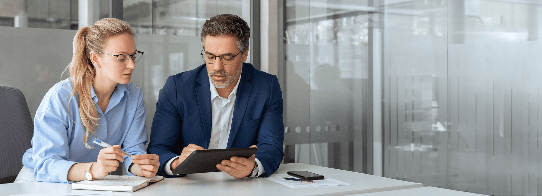 Two business professionals sitting at a desk in a modern office, looking at a tablet together while discussing notes, with a notebook, pen, and smartphone on the table